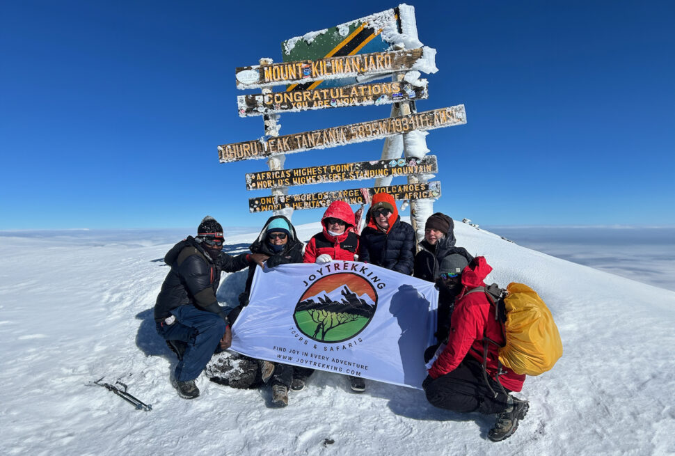 Students celebrating at Mount Kilimanjaro summit in 2024 with JoyTrekking Tours & Safaris flag, showcasing safe and guided trekking experience aligned with Kilimanjaro Safety Essentials