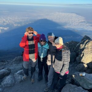 Family of four celebrating at the Mount Meru summit with the mountain’s triangular shadow stretching over the cloud deck, Arusha National Park, Tanzania