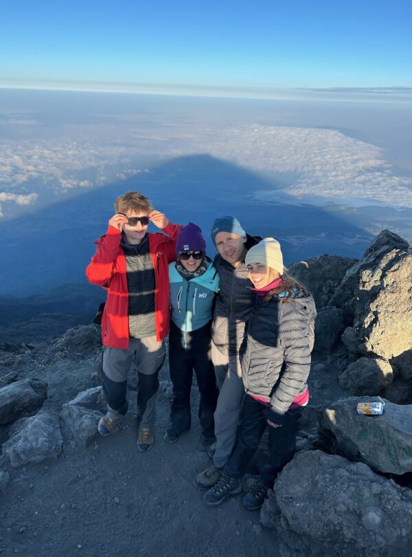 Family of four celebrating at the Mount Meru summit with the mountain’s triangular shadow stretching over the cloud deck, Arusha National Park, Tanzania