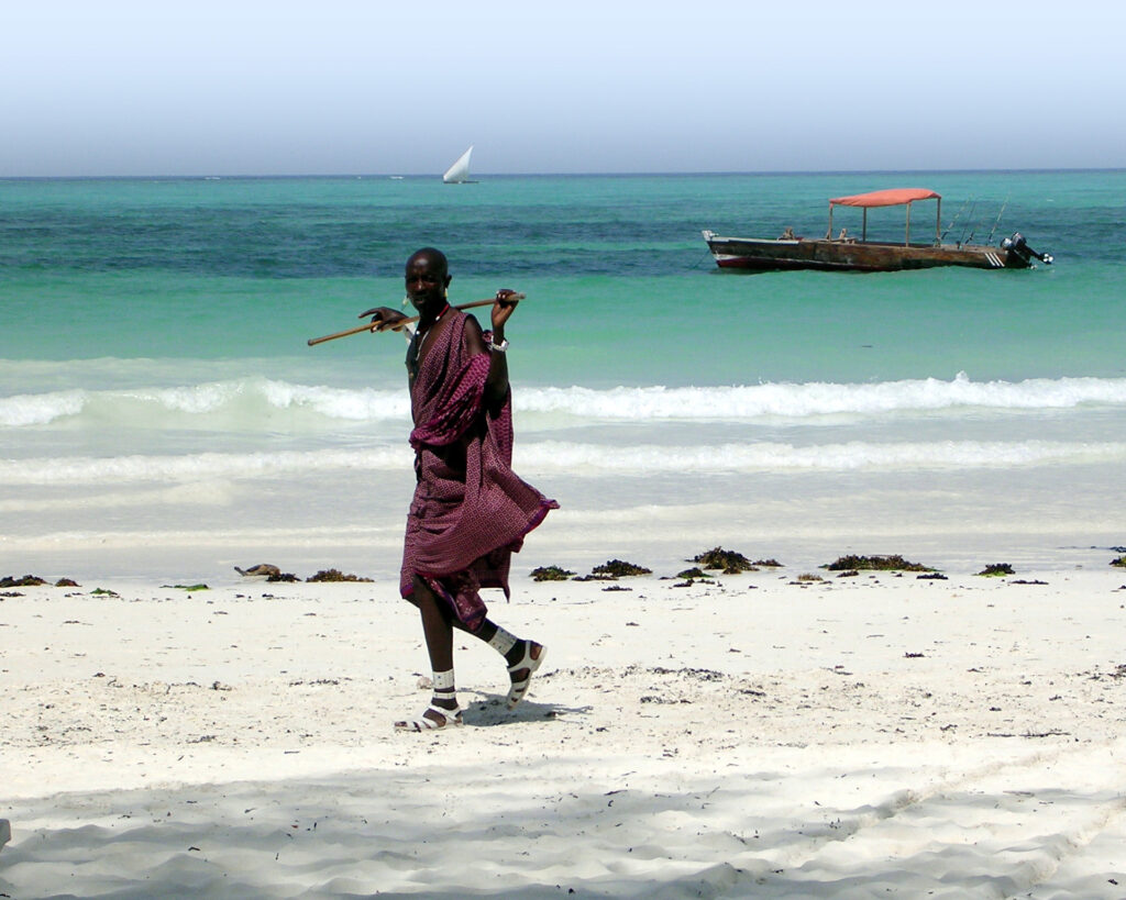 Masaai walking along beach with traditional dhow boat in the background