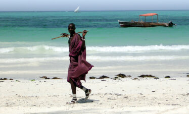 Masaai walking along beach with traditional dhow boat in the background