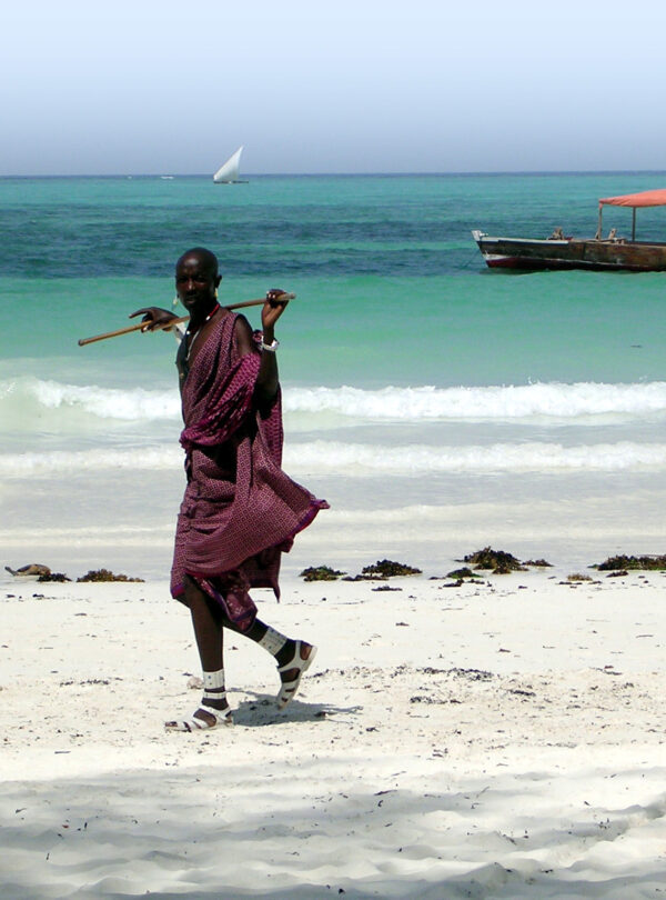 Masaai walking along beach with traditional dhow boat in the background