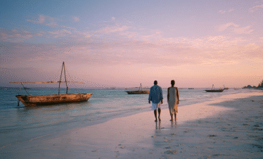 A couple walking barefoot along a Zanzibar beach at sunset, with traditional wooden fishing boats anchored offshore in calm Indian Ocean waters.