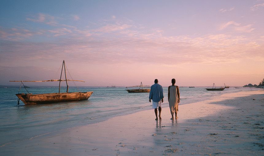 A couple walking barefoot along a Zanzibar beach at sunset, with traditional wooden fishing boats anchored offshore in calm Indian Ocean waters.