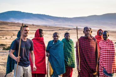 Maasai tribespeople smiling and interacting with an international tourist during a Tanzanian safari, illustrating how safari tourism supports local communities in Tanzania through cultural exchange and community engagement.