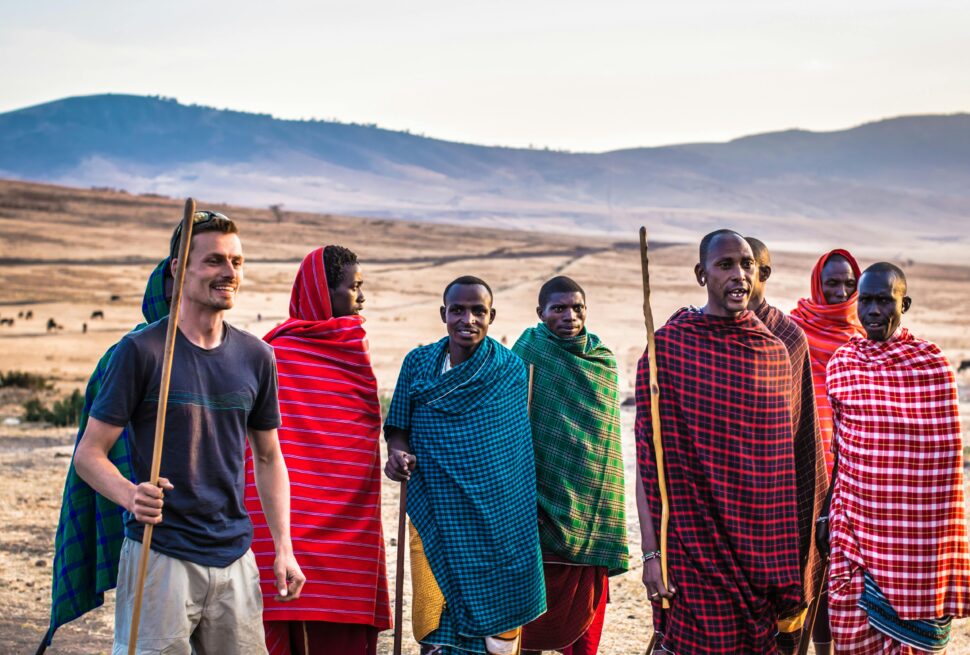 Maasai tribespeople smiling and interacting with an international tourist during a Tanzanian safari, illustrating how safari tourism supports local communities in Tanzania through cultural exchange and community engagement.