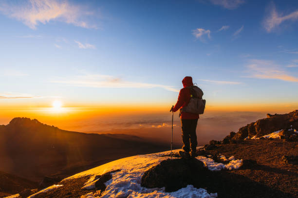 Man with backpack and hiking poles reaches the summit of Mount Kilimanjaro, enjoying a sunrise over Mount Meru during adventure activities in Tanzania