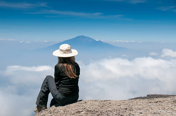 Solo female traveler sitting on Mount Kilimanjaro overlooking clouds and Mount Meru in Tanzania