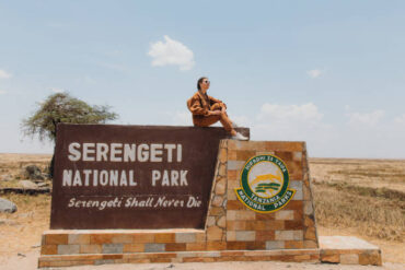 Young woman explorer in safari jumpsuit sitting on a sign overlooking East African savannah, illustrating Tanzania travel safety tips