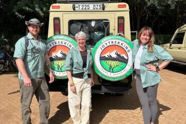 Three international travelers posing beside a safari vehicle branded JoyTrekking Tours & Safaris, representing Tanzania Safari Costs and safari travel experience in Tanzania