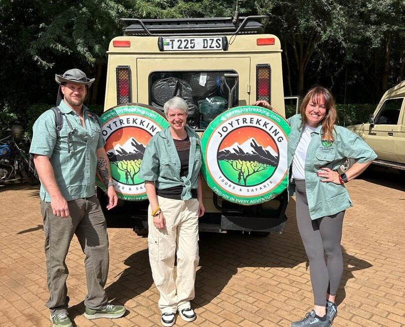 Three international travelers posing beside a safari vehicle branded JoyTrekking Tours & Safaris, representing Tanzania Safari Costs and safari travel experience in Tanzania