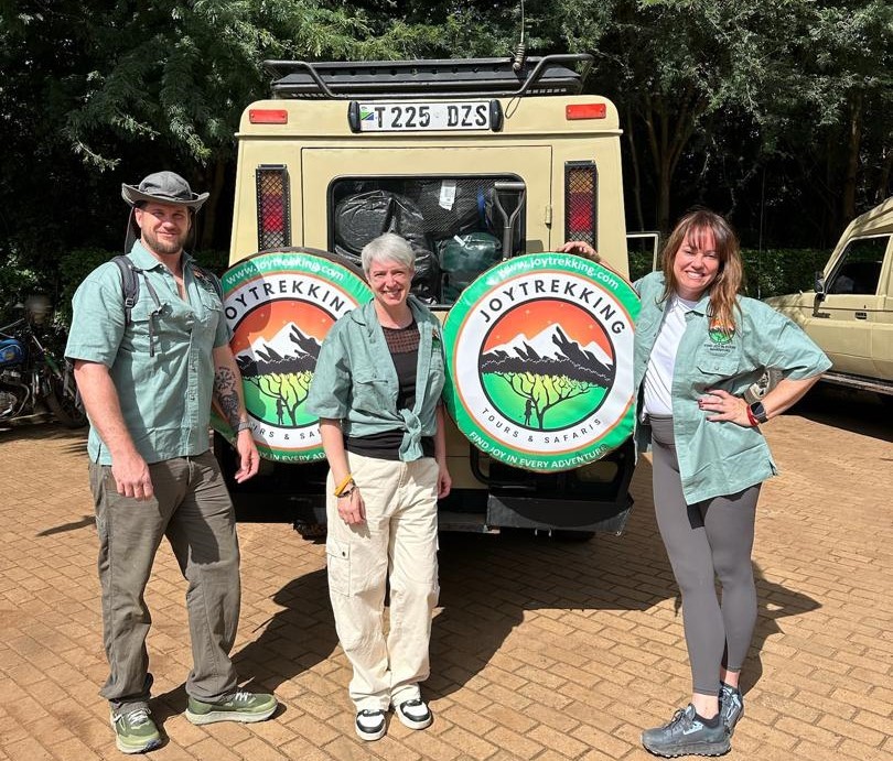 Three international travelers posing beside a safari vehicle branded JoyTrekking Tours & Safaris, representing Tanzania Safari Costs and safari travel experience in Tanzania