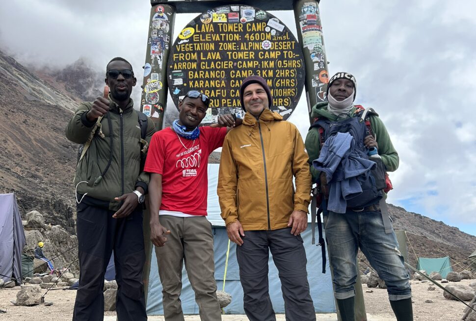 Kilimanjaro Climbing Costs 2026 porter team with guide January Mziray and international traveler at Lava Tower sign during Mount Kilimanjaro trek
