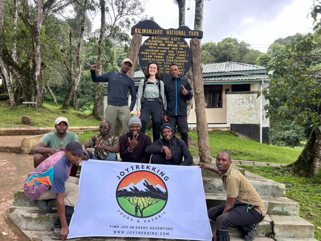 Female tourist Pauline with porters and guide January Mziray holding JoyTrekking Tours & Safaris banner at Machame Gate, responsible tour company in Tanzania Kilimanjaro trek experience