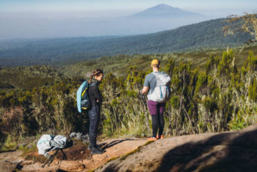 Couple relaxing after hiking Mount Kilimanjaro with backpacks overlooking blue mountain and green valley, representing Kilimanjaro Climbing Cost For Couple adventure experience in Tanzania
