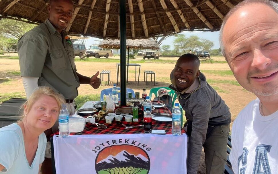 Travelers enjoying lunch with their safari guide during a 5 Day Tanzania Safari Cost experience, with safari Land Cruisers parked inside a national park