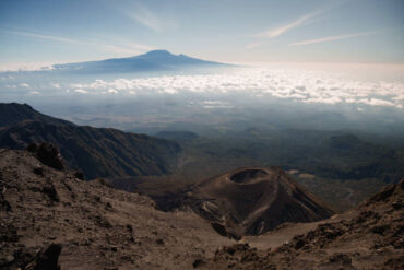 13-Day Mount Meru & Kilimanjaro Combo Cost – Mount Meru and Kilimanjaro, Mount Meru crater with Mount Kilimanjaro in the background under a cloudy sky, Tanzania