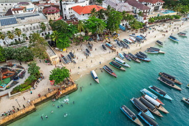 Zanzibar Marine Activities Cost guide featuring Stone Town waterfront with traditional dhow boats, turquoise ocean, and visitors enjoying the beach